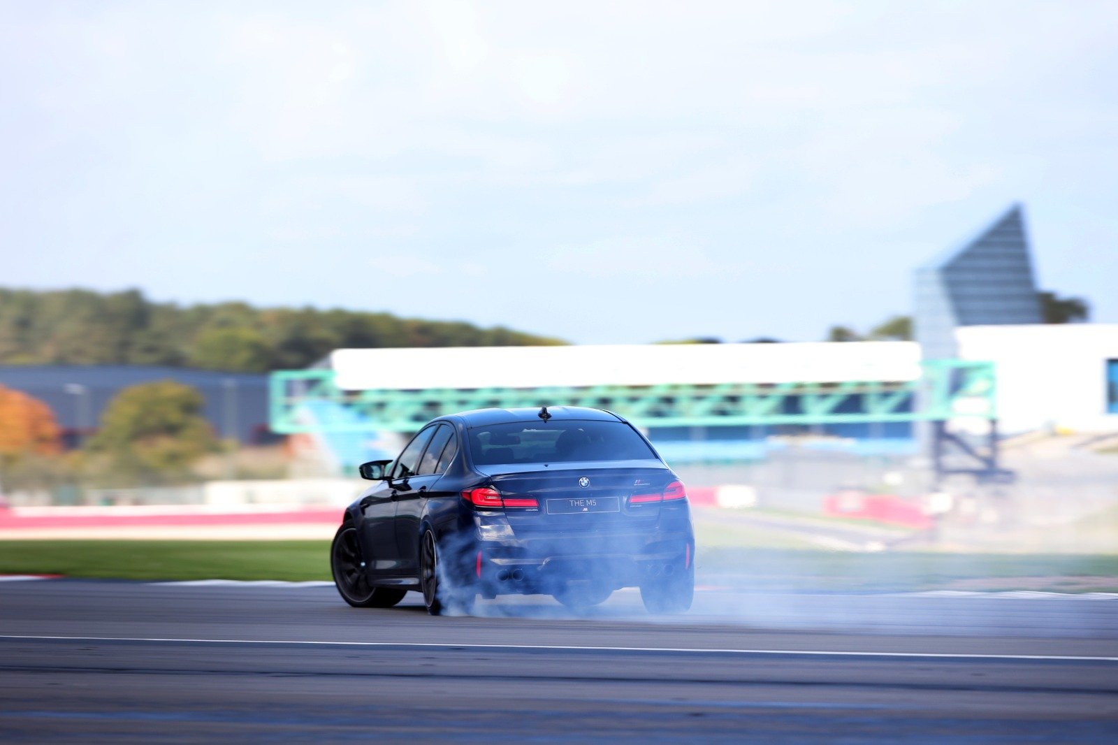 A black BMW M5 Drifting at Silverstone, with smoke coming from the rear tires and blurred background showing a race facility and trees.