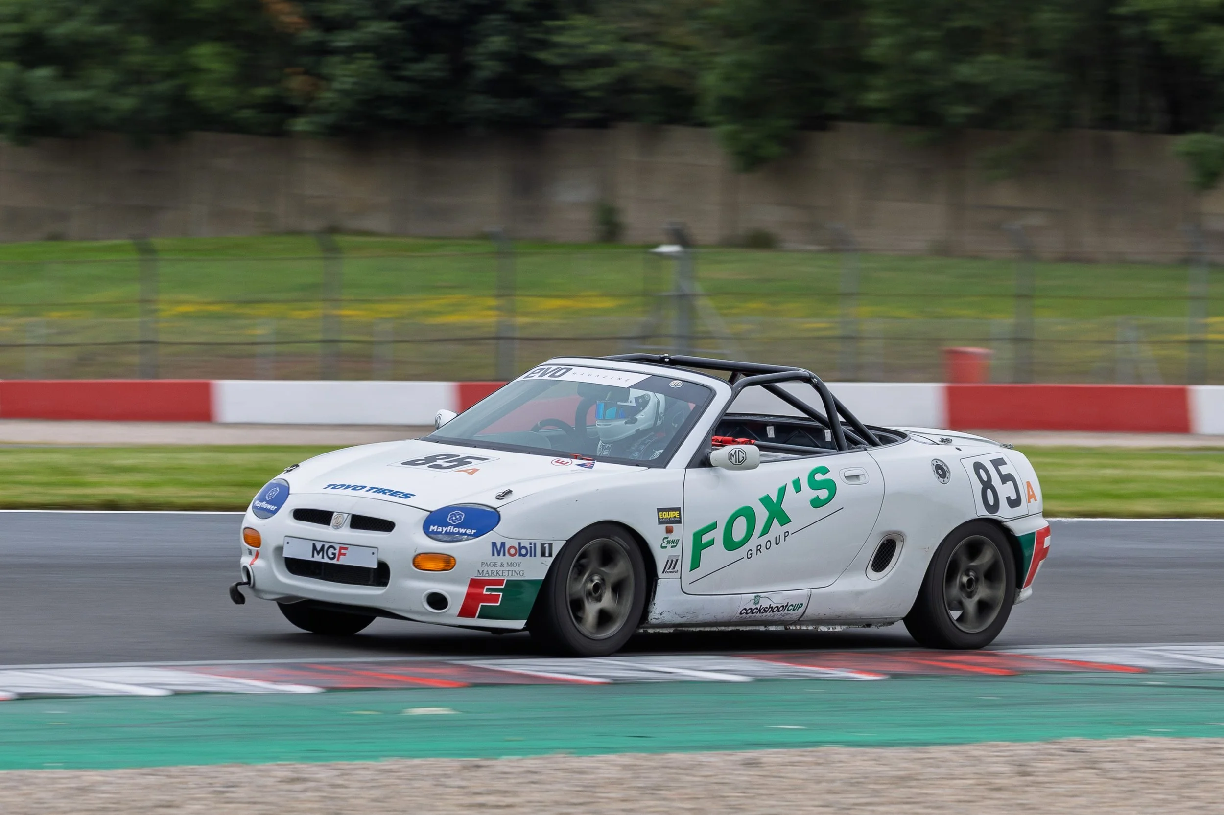 A white race car with the number 85 on its side, moving on a racetrack with red and white barriers and green grass in the background.
