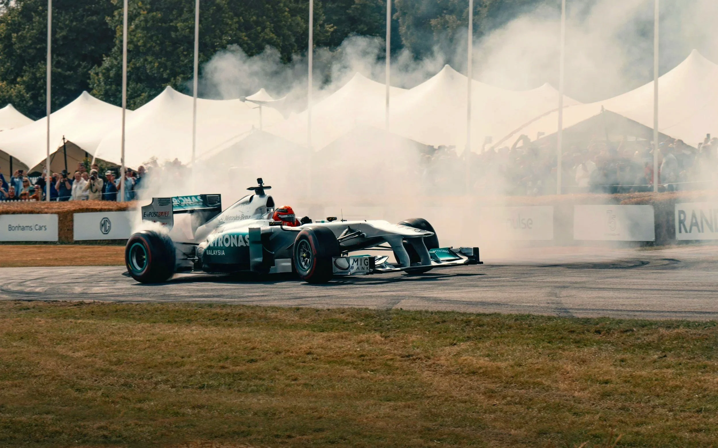 Mercedes F1 car skidding on a track, creating smoke, with spectators and white tents in the background.