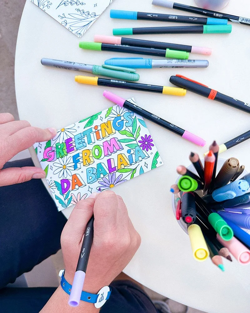 Person coloring a greeting card that says 'Greetings from Da Balia' with colorful markers on a white table.