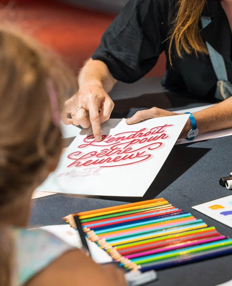 A person is sitting at a table, pointing at a paper with red cursive writing that says "Detroit, we're here." Another person is sitting across from them with colorful colored pencils on the table.