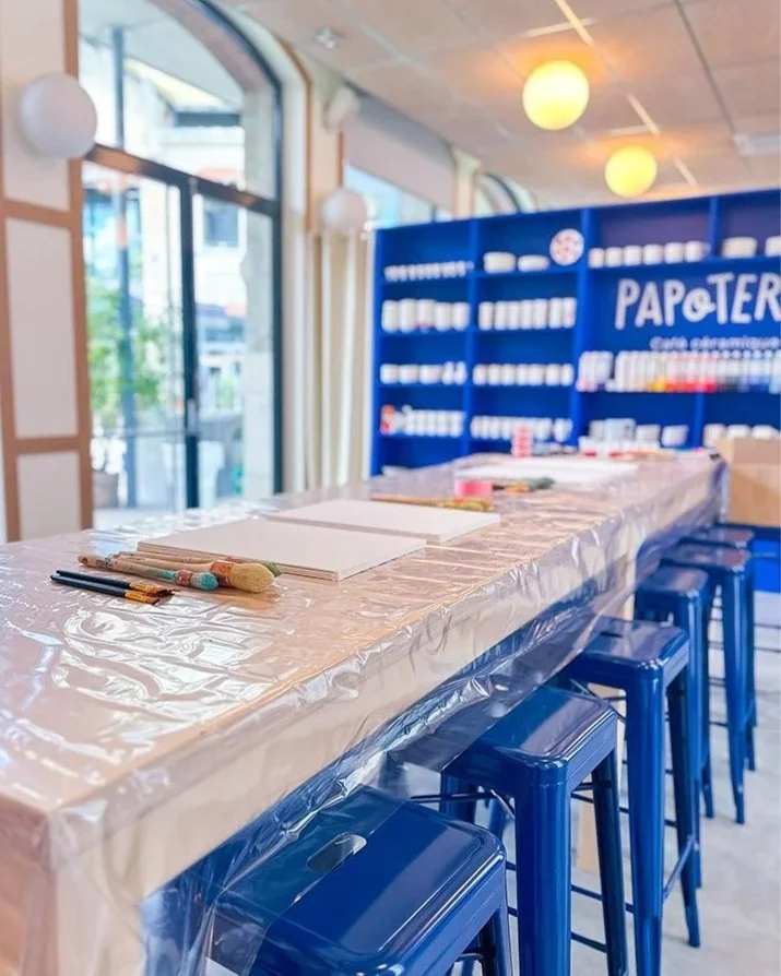 A long table covered with a plastic tablecloth and set up with notebooks, pens, and other art supplies inside a brightly lit cafe or store with large windows. Blue stools are lined up along the table, and a blue shelving unit with white items is visible in the background.