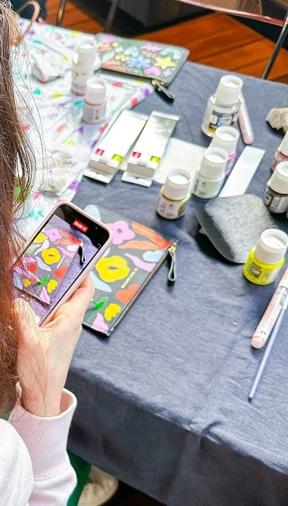 Person taking a photo of a colorful floral painted phone case on a table surrounded by nail polish bottles, a makeup sponge, and art supplies.