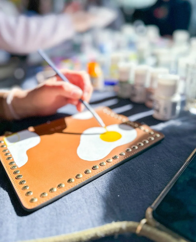 Close-up of a person painting an abstract fried egg on a small canvas, with paint bottles in the background.