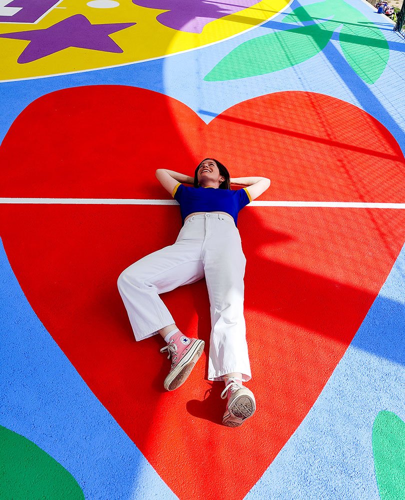 Amy Jones lying on a colorful outdoor basketball court that has a large red heart painted on it, with her arms behind her head and smiling.