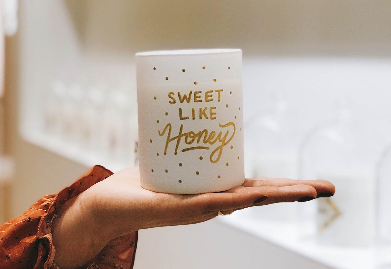 A hand holding a white mug with gold lettering that says 'Sweet like honey' and gold polka dots, with a blurred background.