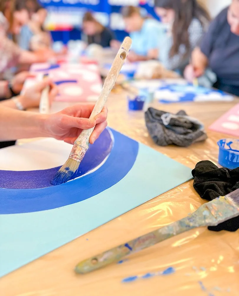 A person painting a circular blue design on paper with a paintbrush, with a group of people in the background in an art class.