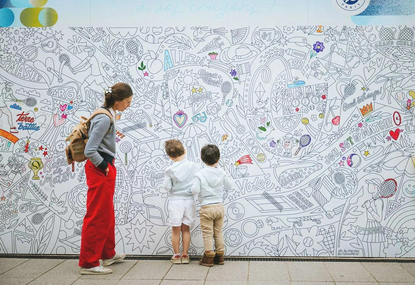 A woman and two young boys are looking at a large, white mural filled with colorful doodles and words related to tennis and sports.