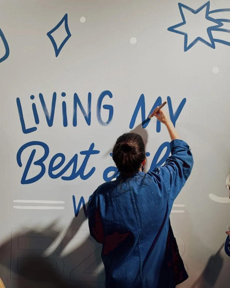 A person with dark hair tied back, wearing a blue jacket, is writing on a white wall with a blue marker. The wall has the words 'LIVING MY Best' written on it, along with decorative stars and dots.