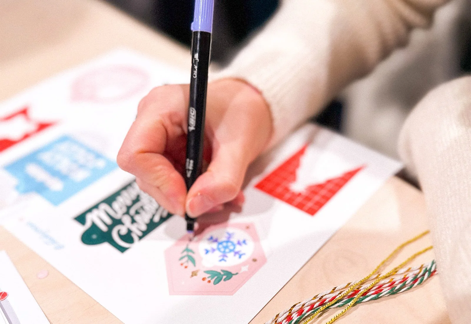 A person is writing a holiday-themed design on a sticker with a purple marker. The design includes a blue snowflake, a pink tag, and green foliage. The surface has other stickers, a red and white patterned design, and some red, green, and gold twine.