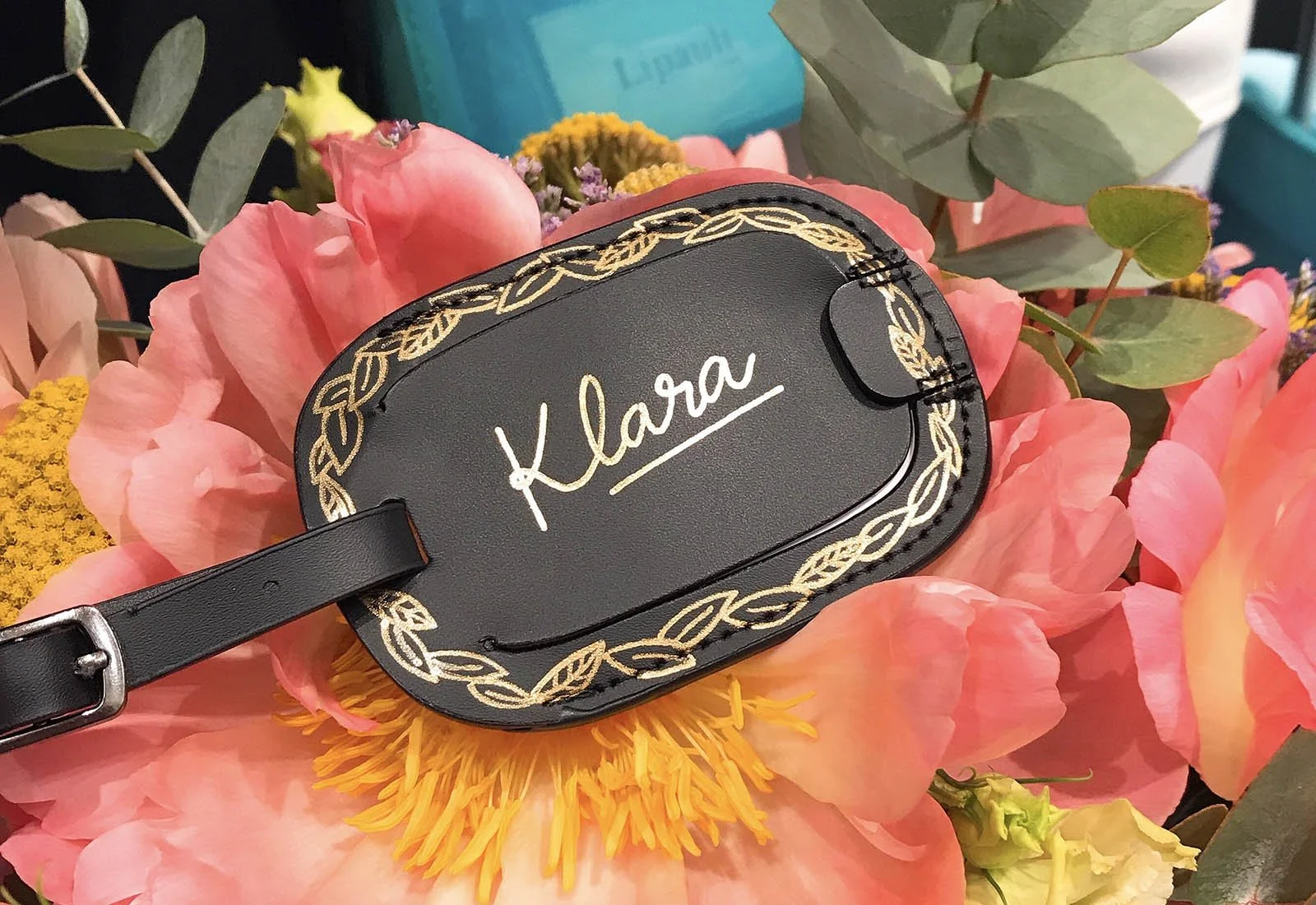 A black leather luggage tag with a gold chain pattern border and the name 'Klara' written in gold script, placed on pink and yellow flowers with green leaves.