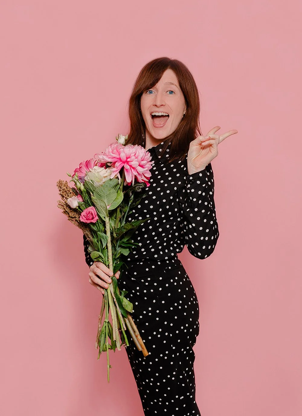 Amy Jones holding a bouquet of pink flowers, wearing a black polka-dot outfit, standing against a pink background, smiling and making a peace sign.
