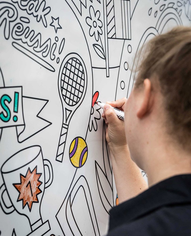 A person coloring a large black-and-white wall mural with various sports and celebration-themed drawings, including tennis rackets, a ball, a cup, and banners.