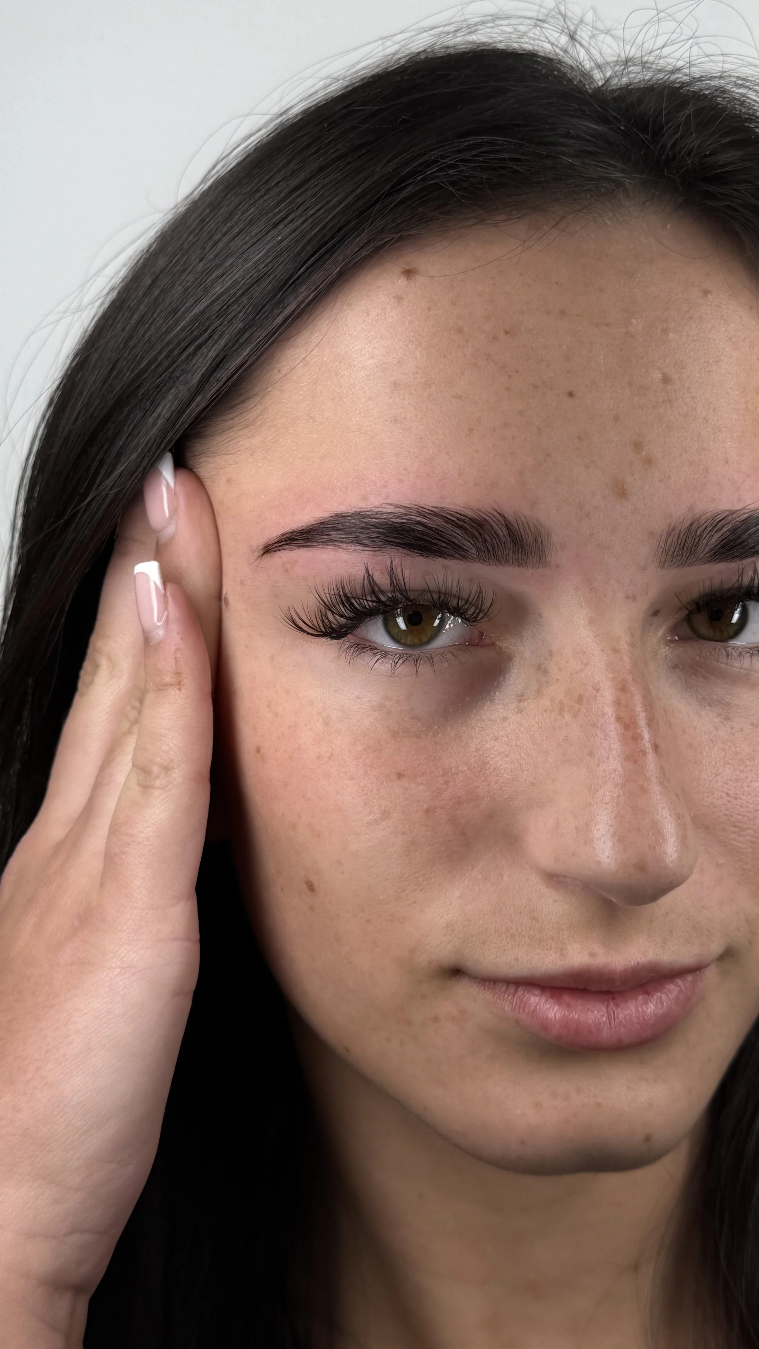 Close-up of a young woman with brown eyes, long dark hair, and natural skin with freckles. She is gently touching her ear with her left hand and has a subtle smile.