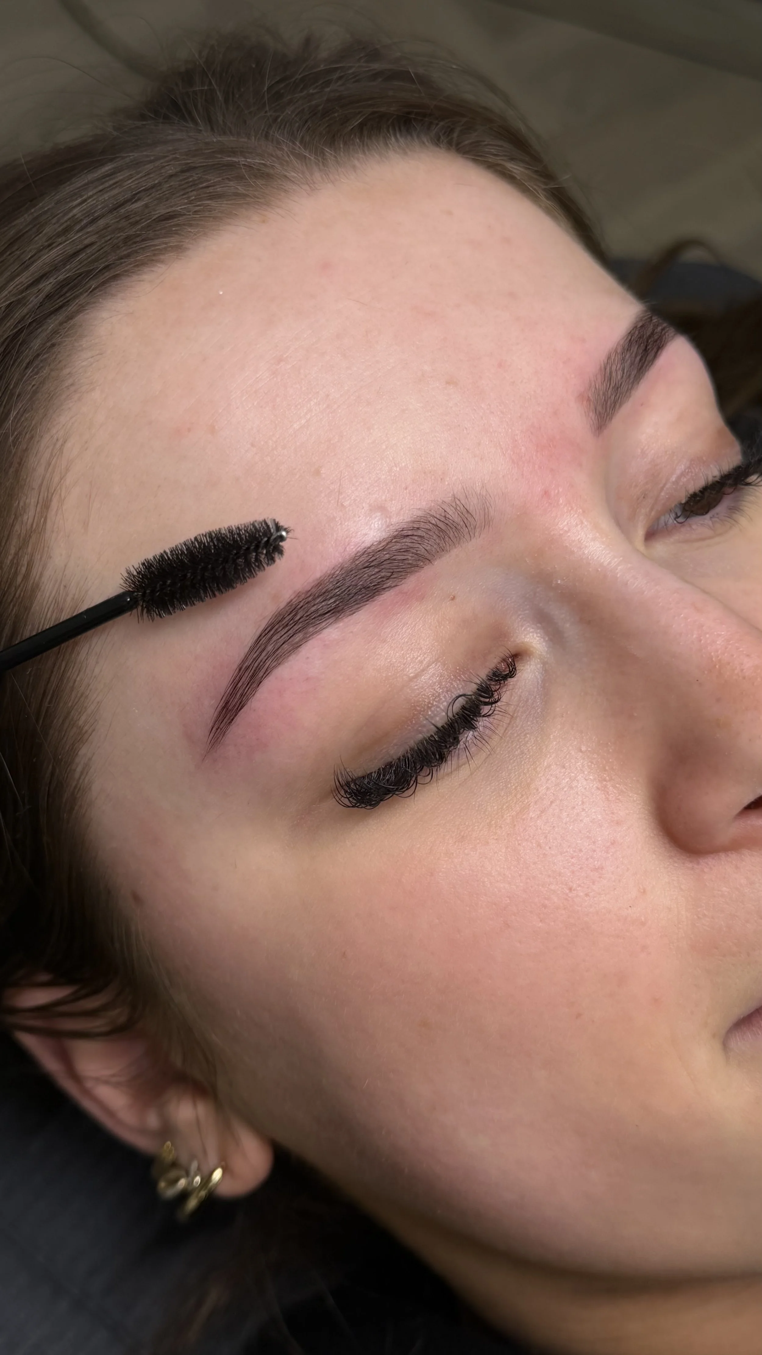 Close-up of a woman receiving eyelash extension application, with one eye closed and a black mascara wand near her eyebrow.