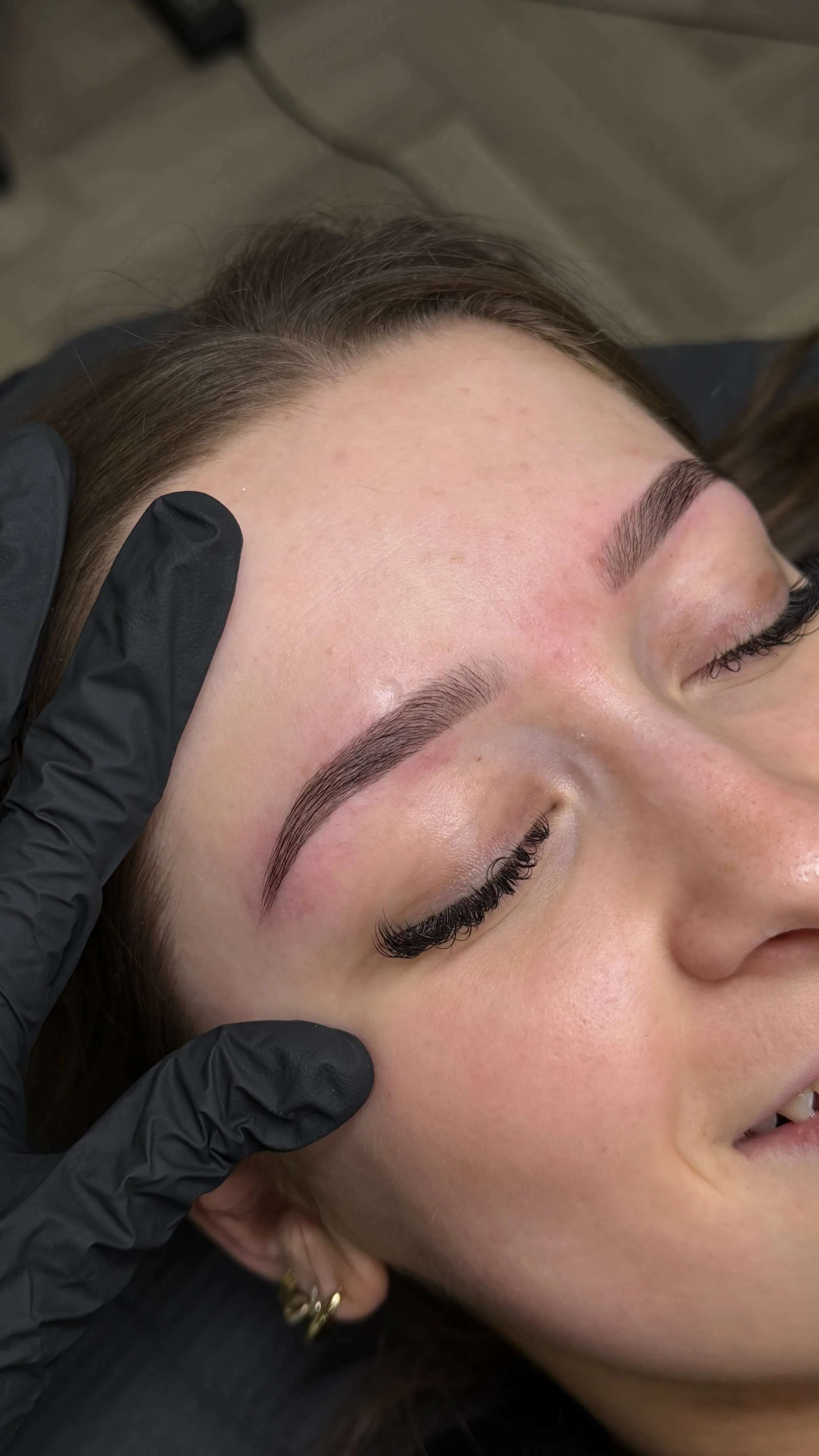 Close-up of a woman with newly microbladed eyebrows, lying down with eyes closed, during a cosmetic procedure in a salon or clinic.
