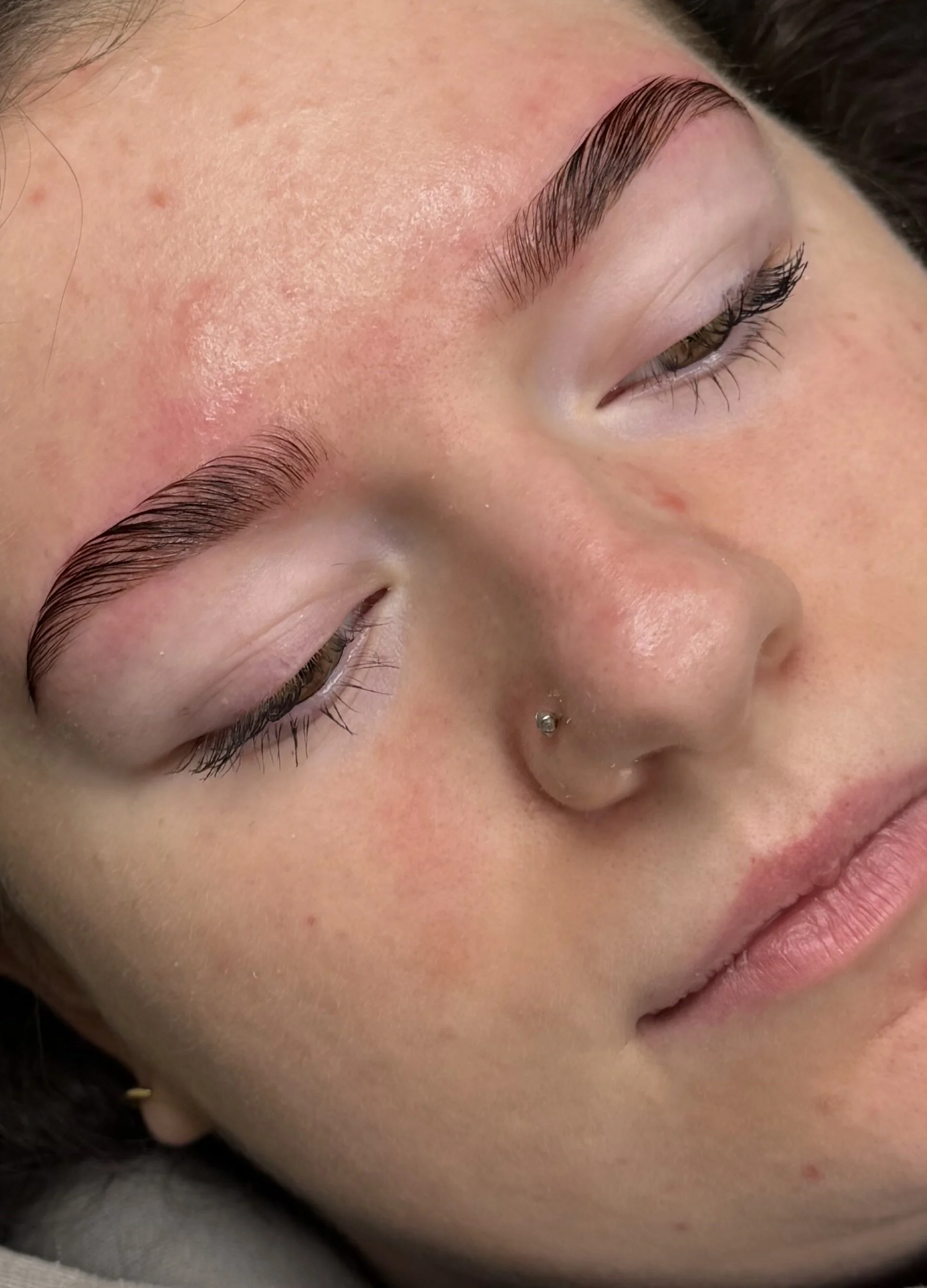 Close-up of a woman's face with well-groomed eyebrows, closed eyes, a nose piercing, and minimal makeup.