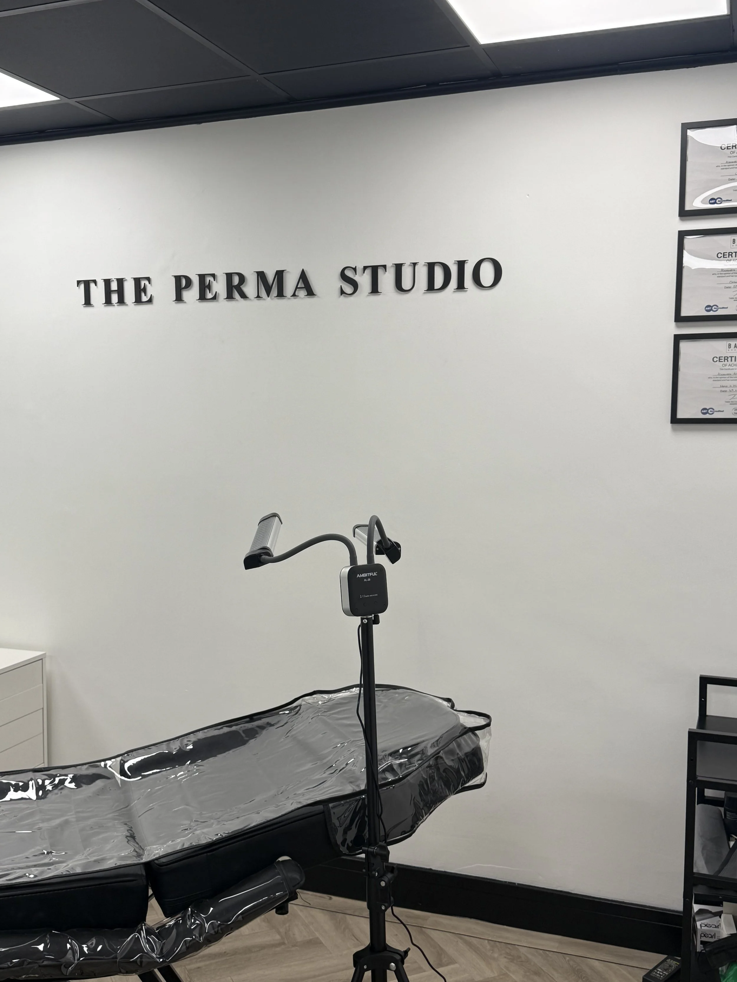 A medical examination room with a treatment table covered in clear plastic, an adjustable lamp, framed certificates on the wall, and a sign that reads 'The Perma Studio'.