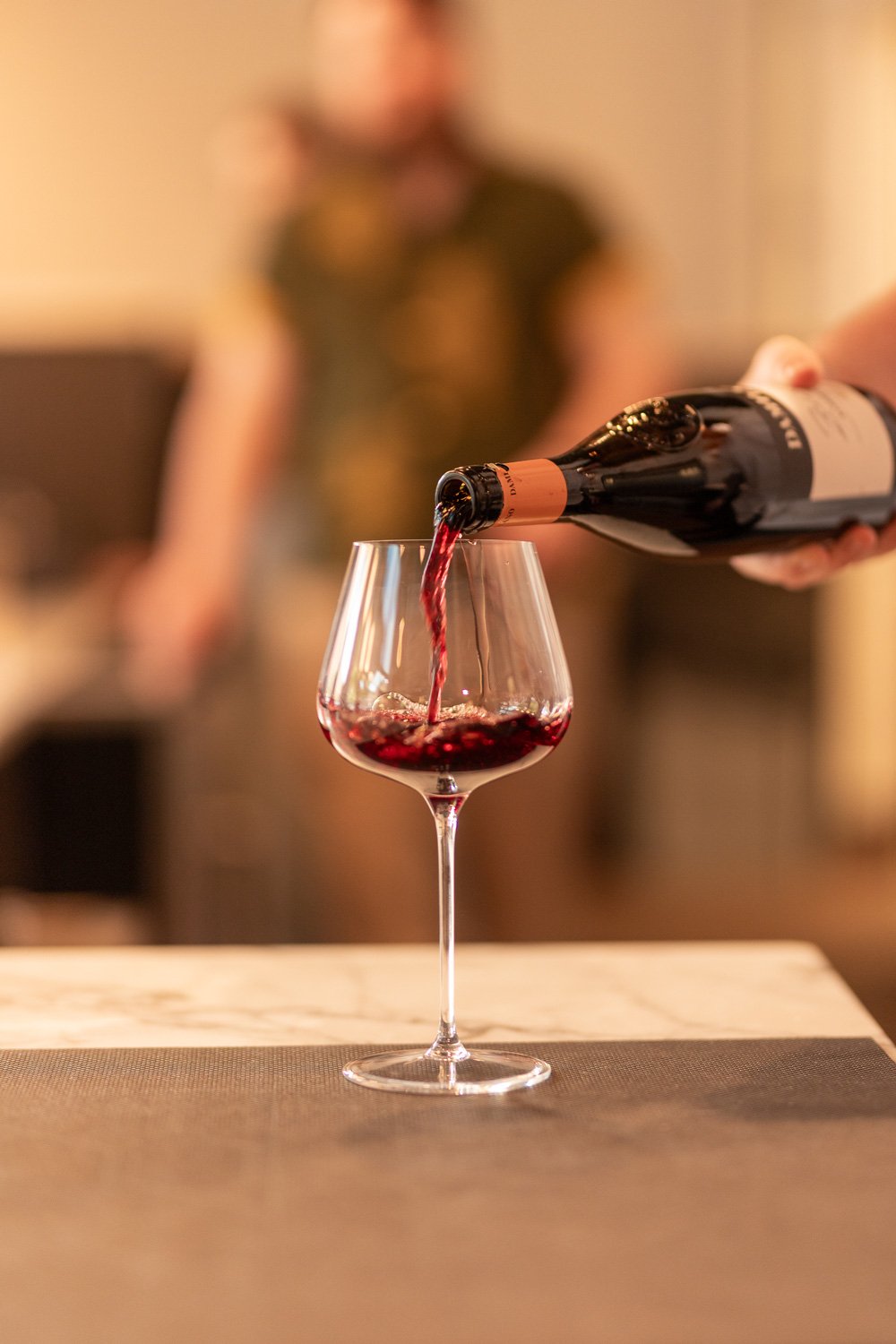 Person pouring red wine into a wine glass on a table with a blurred background.