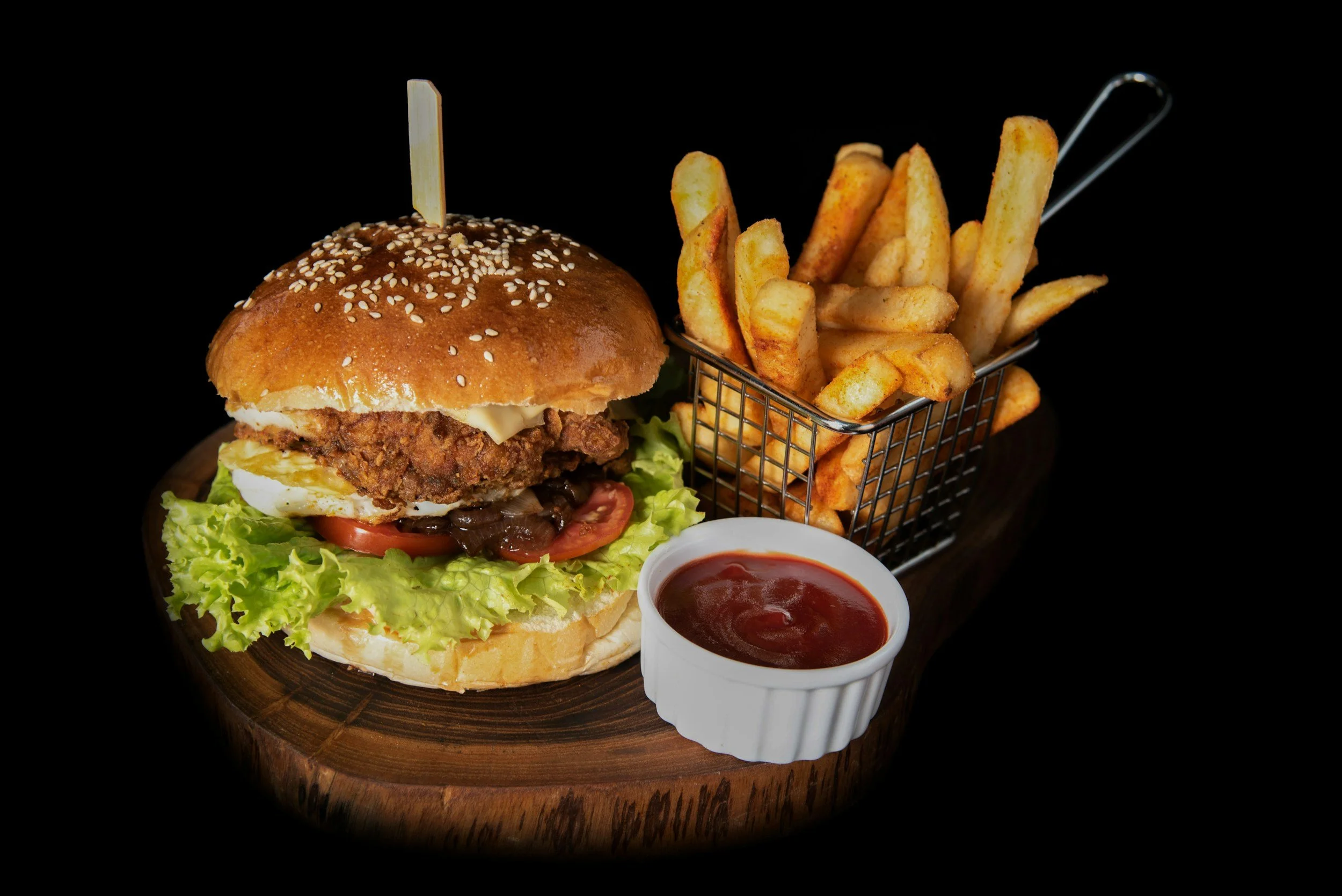 Gourmet burger with fried chicken patty, lettuce, tomato, pickles, cheese, and bun, served with French fries in a metal basket and a side of ketchup on a wooden board.