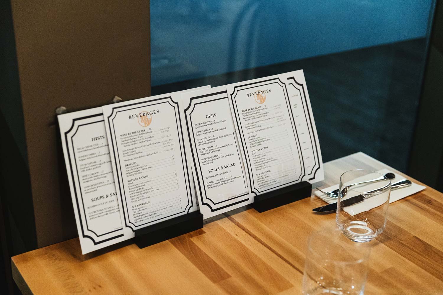 Restaurant table with three black menu stands displaying drinks, starters, soups, salads, and beverage options. Clear drinking glass, fork, knife, and spoon on white napkin on wooden table.