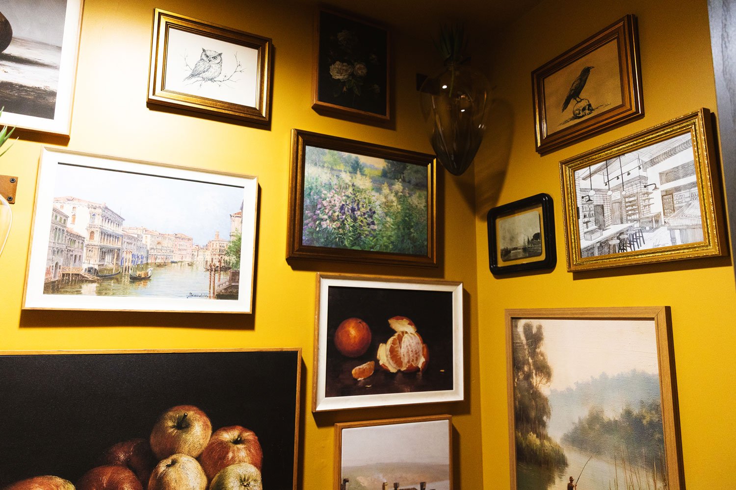 Wall decorated with multiple framed pictures, including paintings and sketches of landscapes, cityscapes, still life, animals, and floral scenes, on a yellow wall.