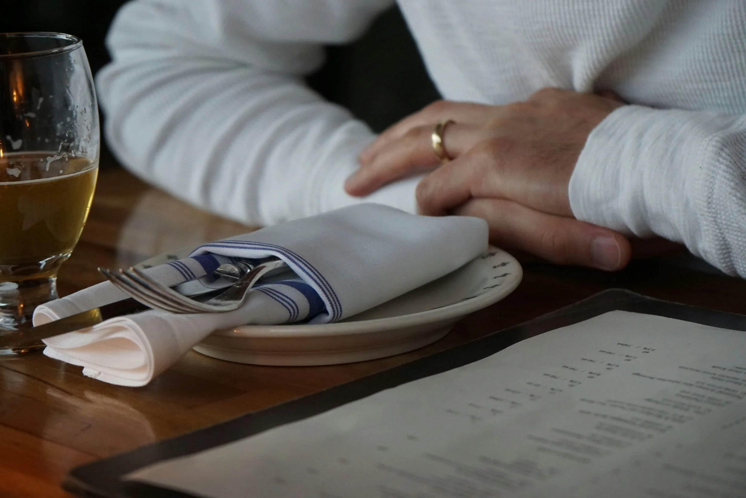 Person holding hands on a restaurant table, with a glass of beer, white cloth napkin, and cutlery on a plate.