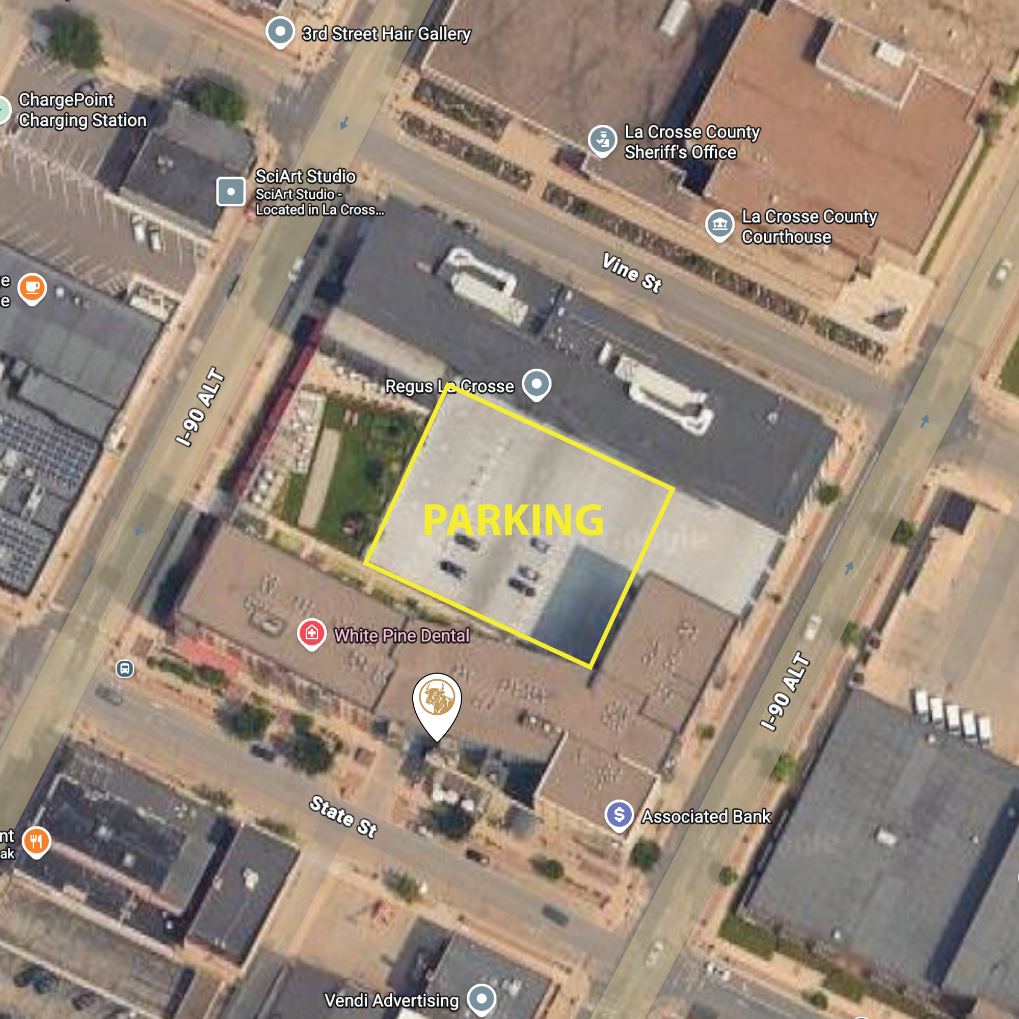 A satellite view of a parking lot outlined in yellow with the word 'PARKING' in large yellow letters in the center. The parking lot is surrounded by streets and buildings, including White Pine Dental, a bank, and various businesses. The parking lot has some cars parked in it.