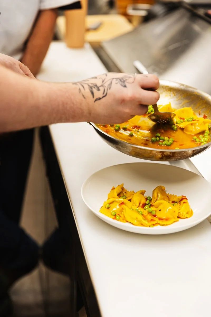 A person with a tattoo on their arm serving yellow tortellini pasta with green peas in a broth from a metal pan onto a white plate.