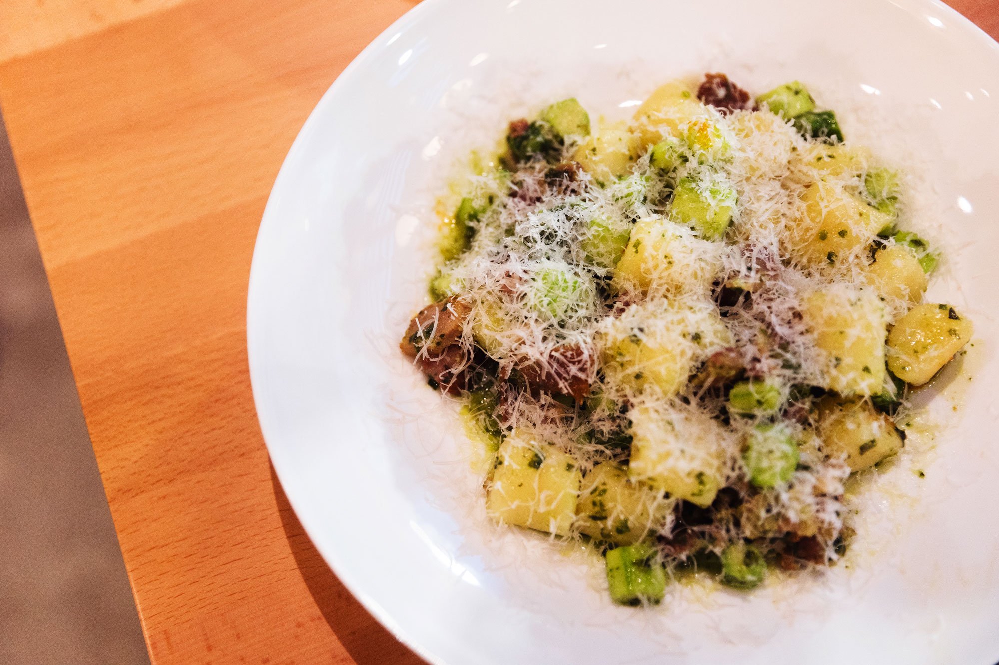 A white bowl of gnocchi with shredded cheese and chopped green onions on a wooden table.