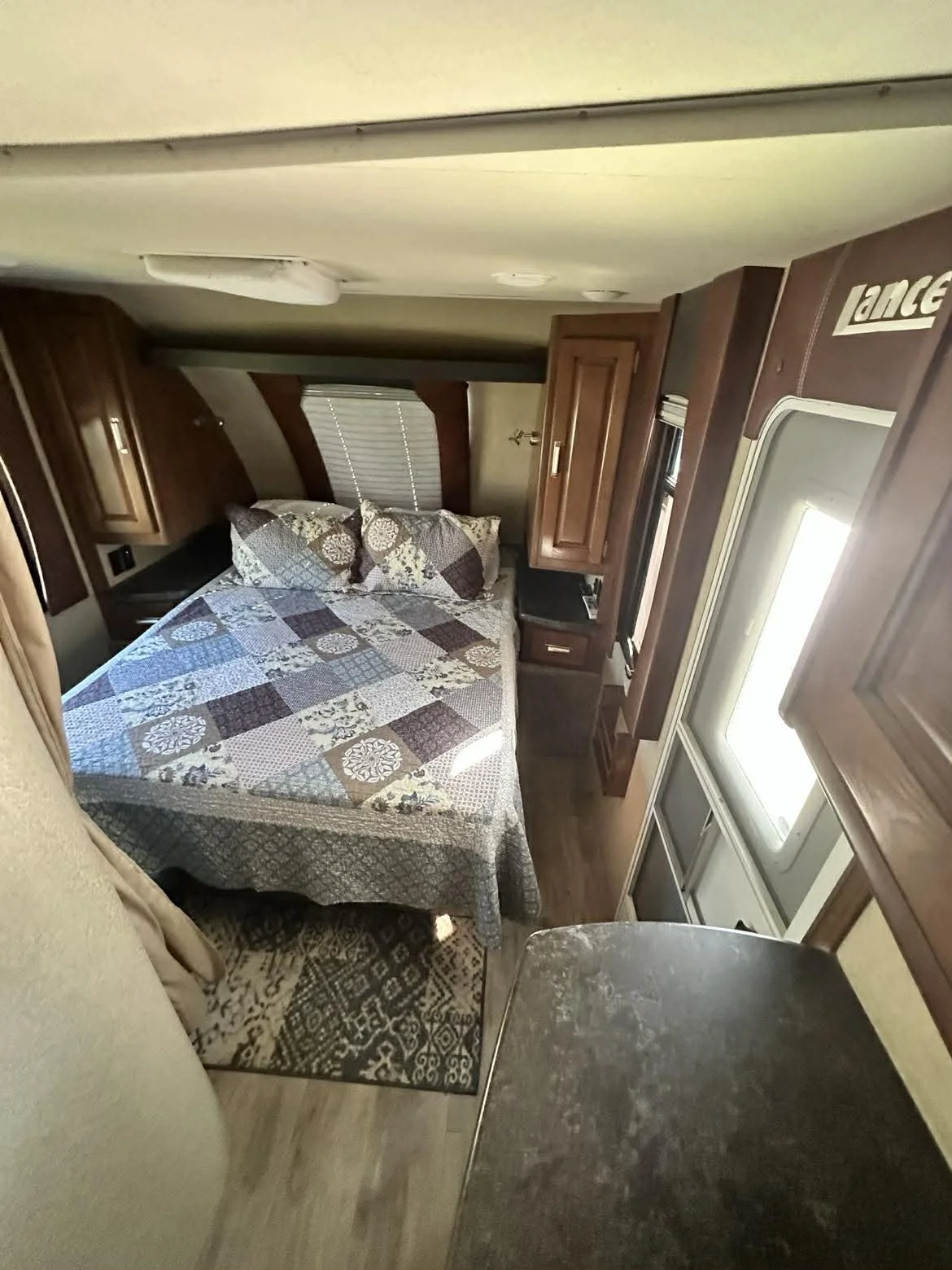 Interior of a compact bedroom inside a camper, featuring a bed with a patterned quilt and pillows, wooden cabinets, a window with blinds, and a small table or countertop.
