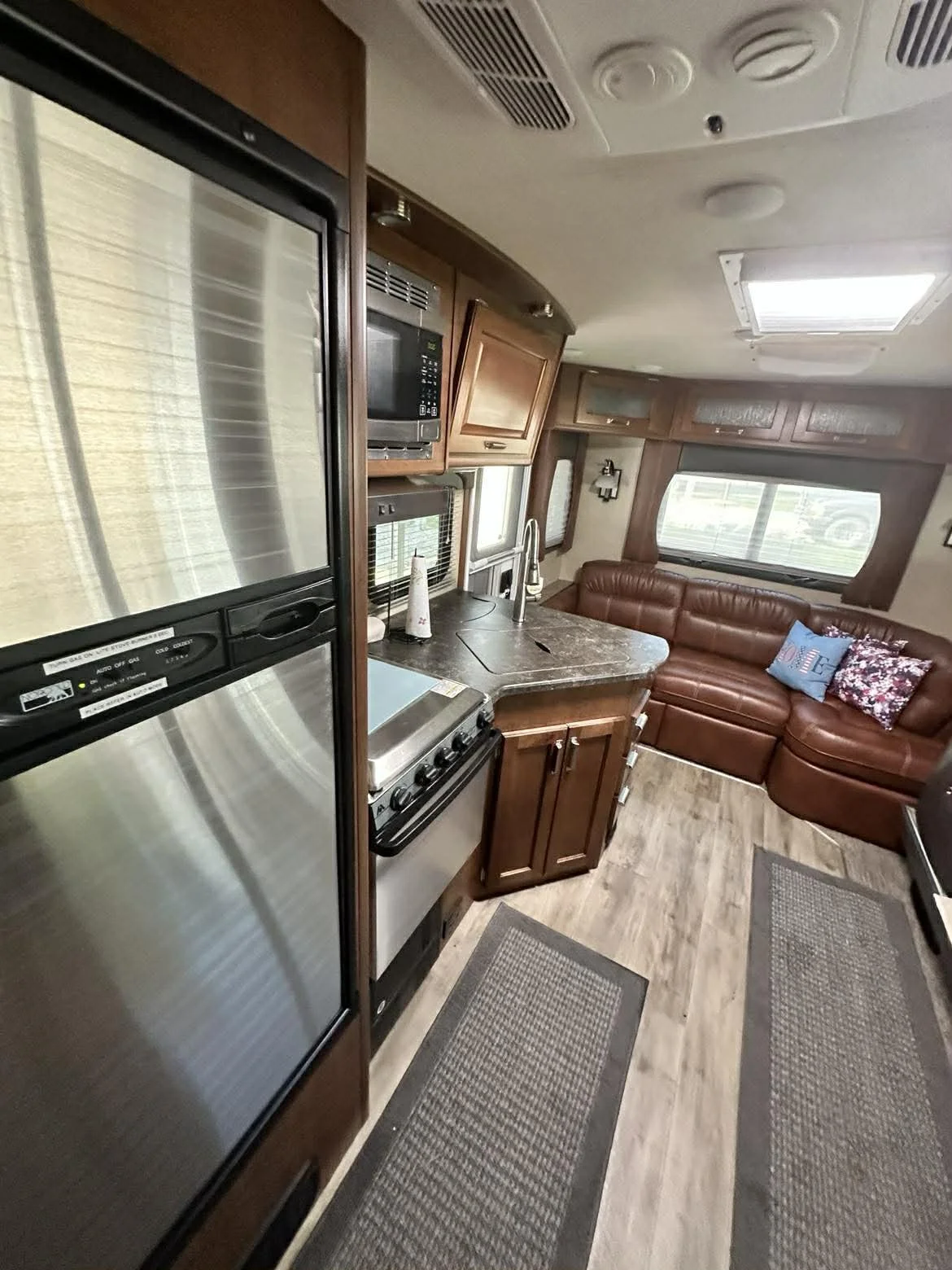 Interior of an RV kitchen with a refrigerator, stove, microwave, and wooden cabinets, with a leather sofa and windows in the background.