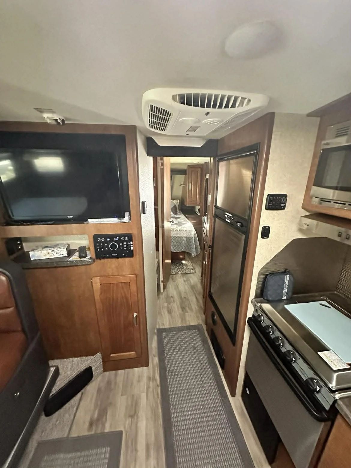 Interior of an RV showing a corridor with wooden cabinetry, a flat-screen TV, a control panel, a kitchen stove, and a refrigerator, with a bedroom visible in the background.