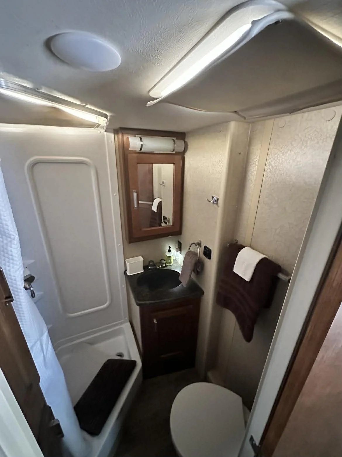 Small bathroom with a shower, mirror, sink with countertop, towel rack with folded towel, toilet, and wood-framed mirror in a compact space, likely in an RV or camper.