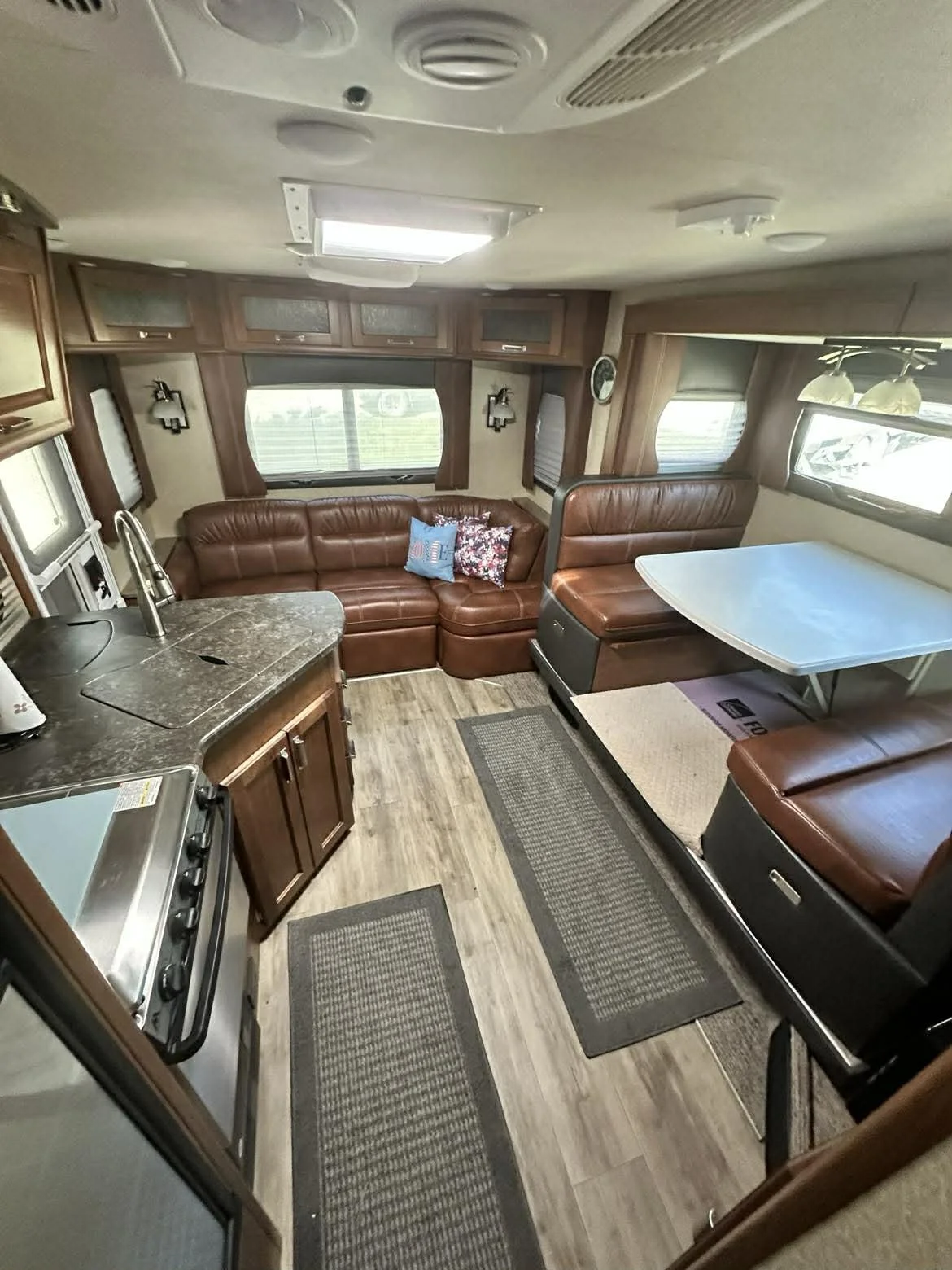 Interior of an RV with a curved leather sofa, kitchen area with stove and sink, wood cabinets, and dining booth with a table. Windows with blinds, ceiling lights, and wood flooring.