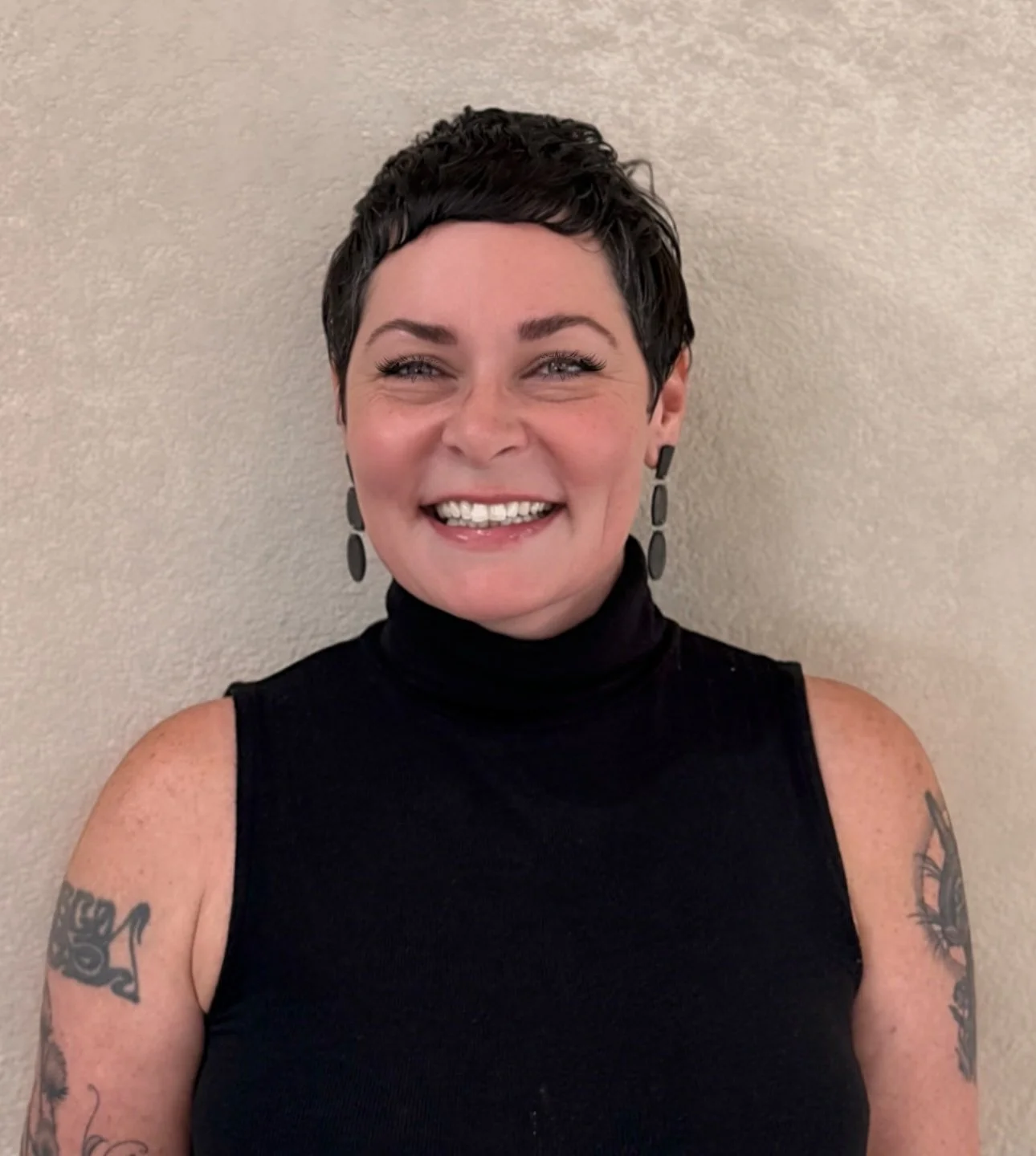 A smiling woman with short dark hair, wearing a black sleeveless turtleneck and black earrings, standing against a beige wall.