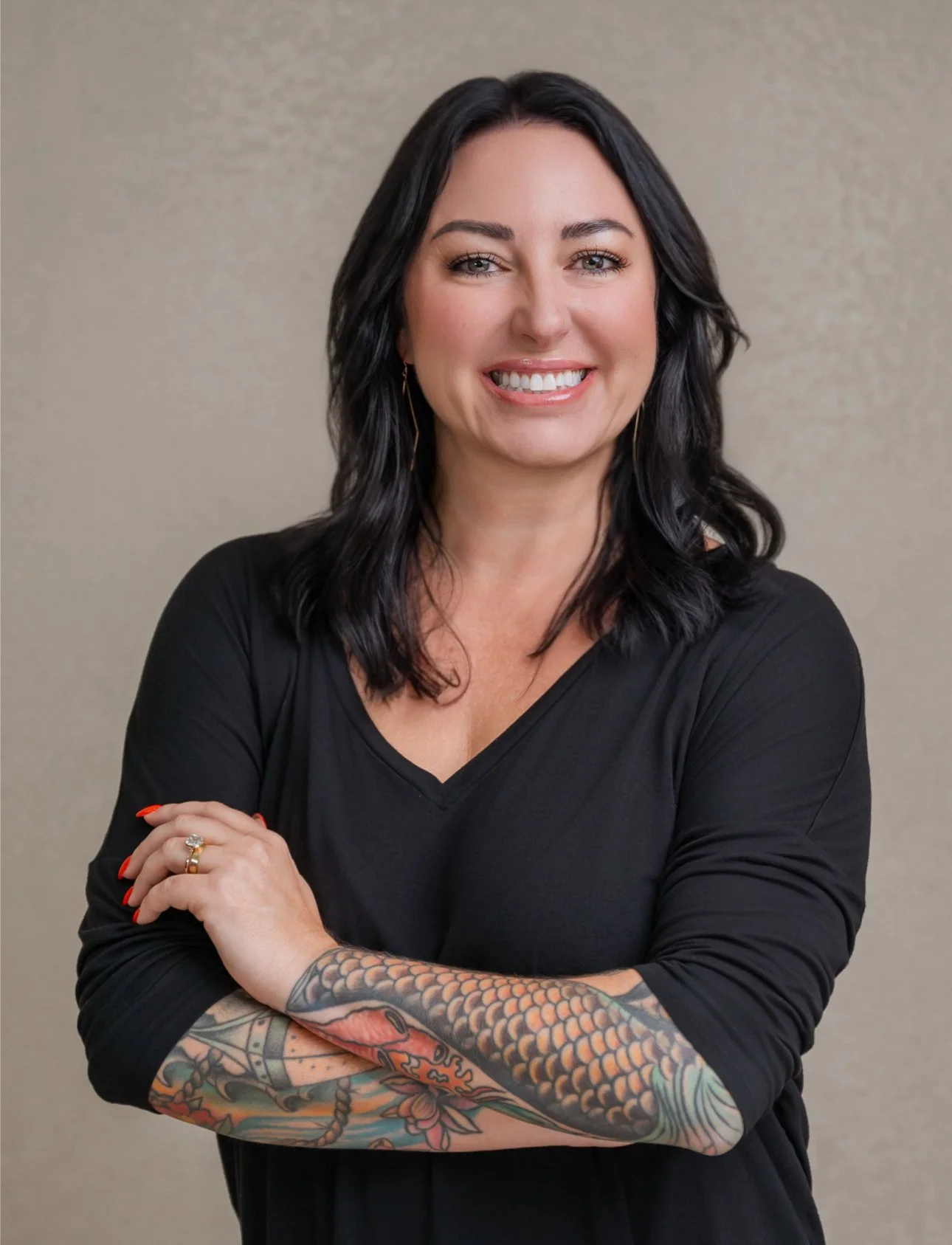 A woman with black hair smiling, wearing a black top, with a tattoo sleeve on her right arm, standing against a plain beige background.