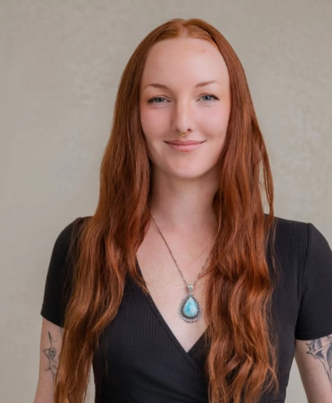 A woman with long red hair and fair skin, wearing a black top, a turquoise pendant necklace, and a septum piercing, standing against a plain background.