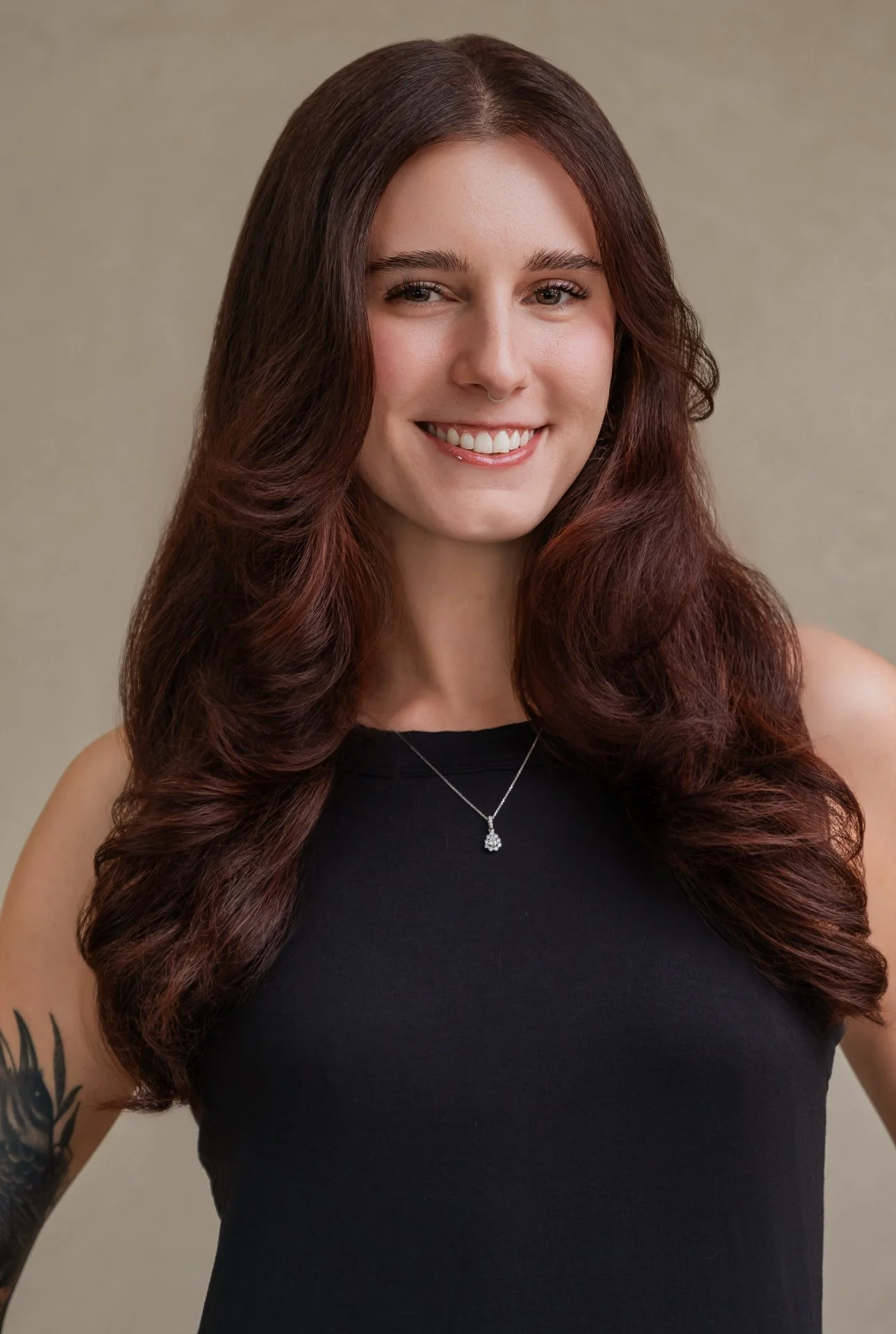 A woman with long, wavy reddish-brown hair smiling at the camera, wearing a black sleeveless top, a necklace with a pendant, and a visible tattoo on her left arm, standing against a plain background.