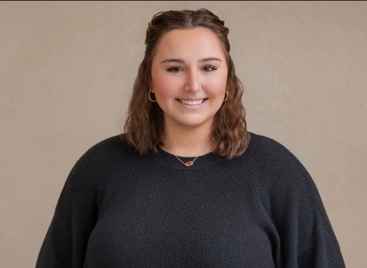Portrait of a young woman with shoulder-length brown curly hair wearing a black sweater, gold hoop earrings, and a necklace with a red pendant, smiling against a plain beige background.