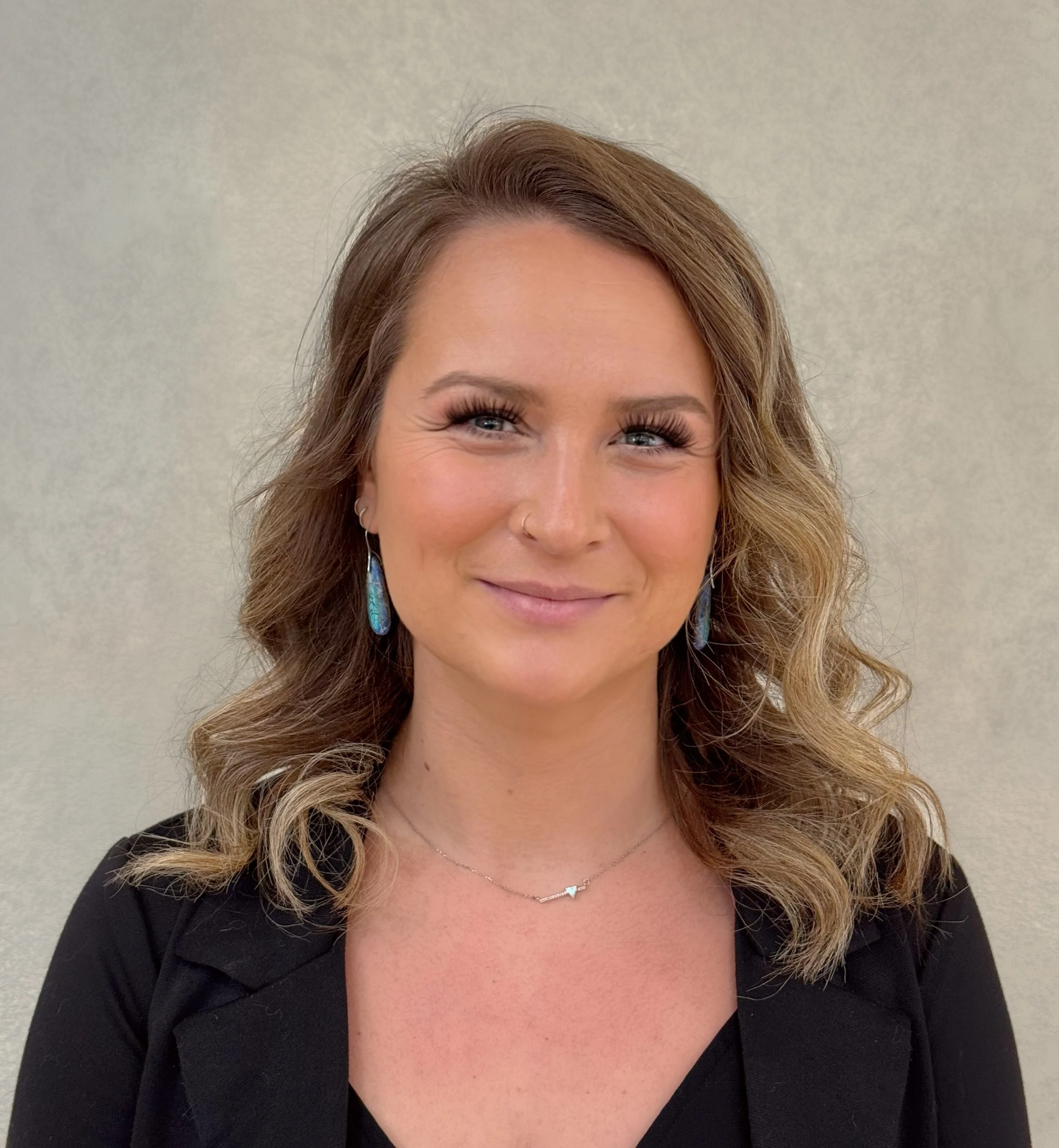 A woman with wavy light brown hair, blue eyes, and a nose piercing smiling at the camera, wearing earrings, a necklace, and a black blazer against a beige wall.