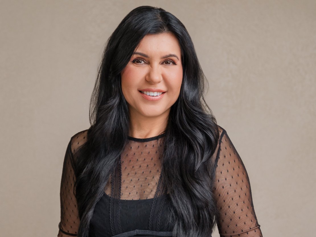 A woman with long black hair, smiling, wearing a black polka-dot sheer top, standing against a beige wall.