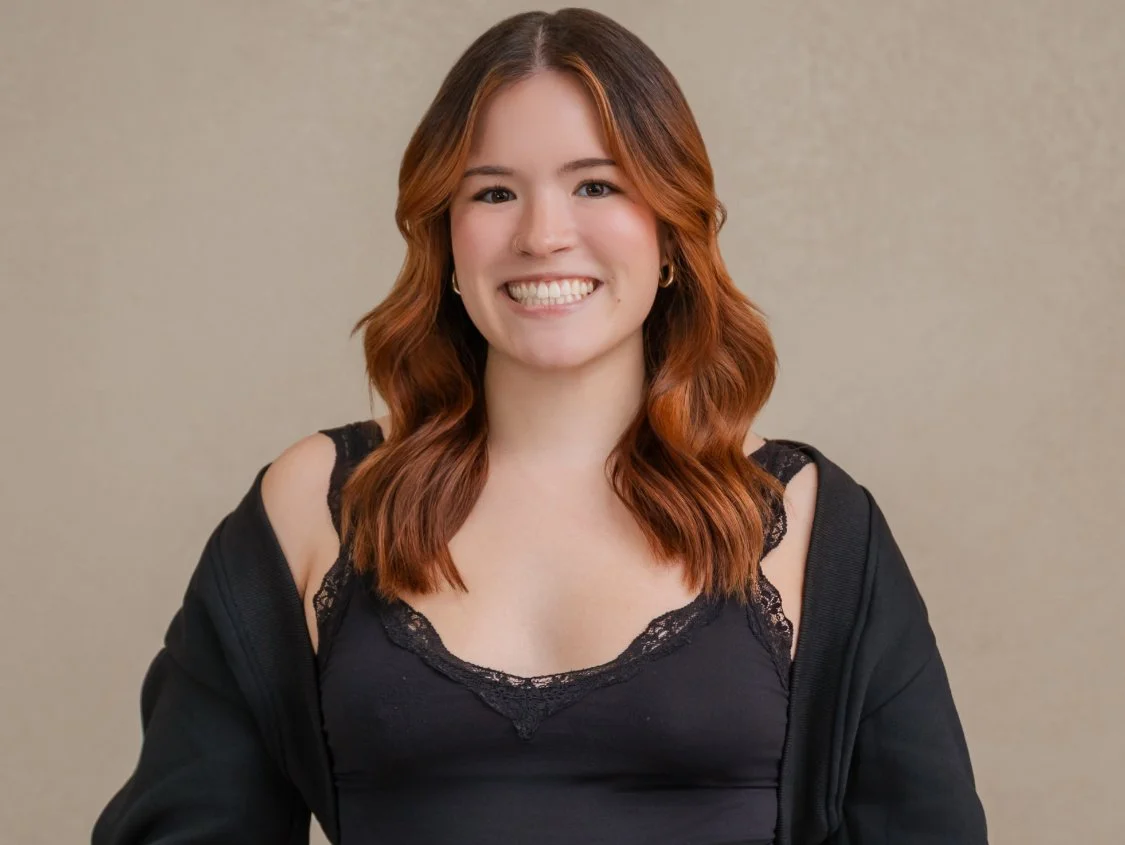 A smiling woman with shoulder-length red hair, wearing a black tank top with lace trim and a black jacket, standing against a neutral background.
