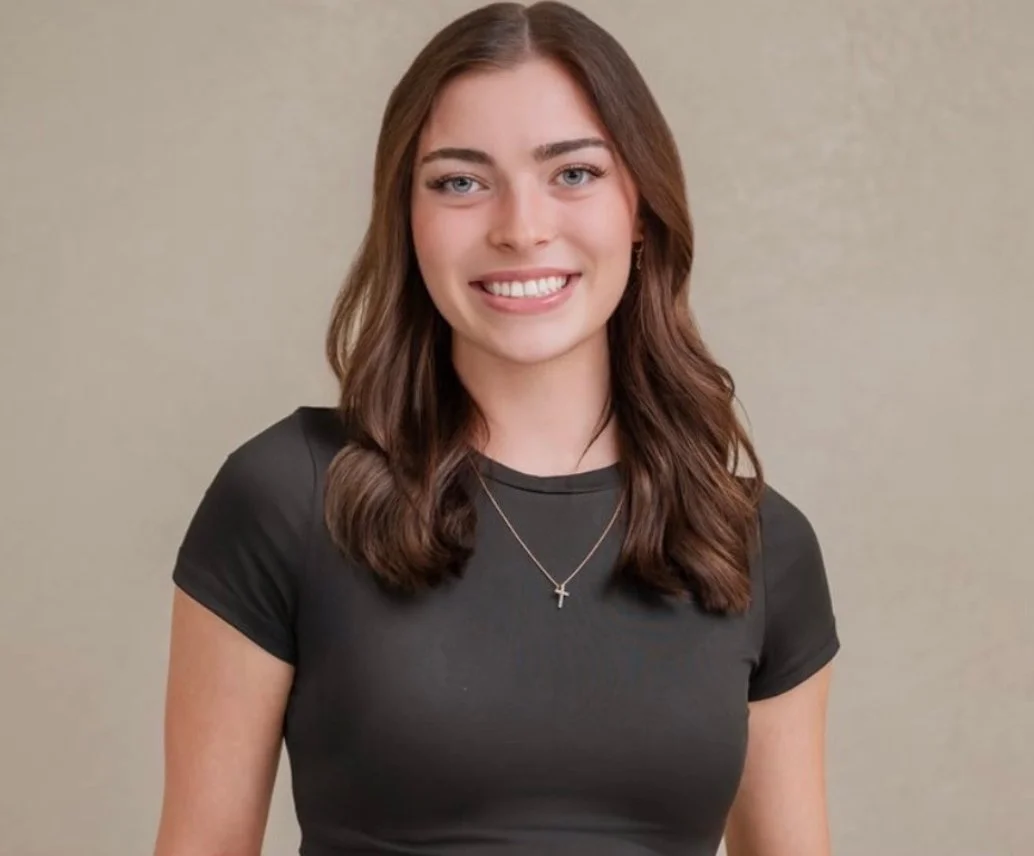 A young woman with shoulder-length brown hair, wearing a black T-shirt and a silver necklace with a cross, smiling at the camera against a plain beige background.