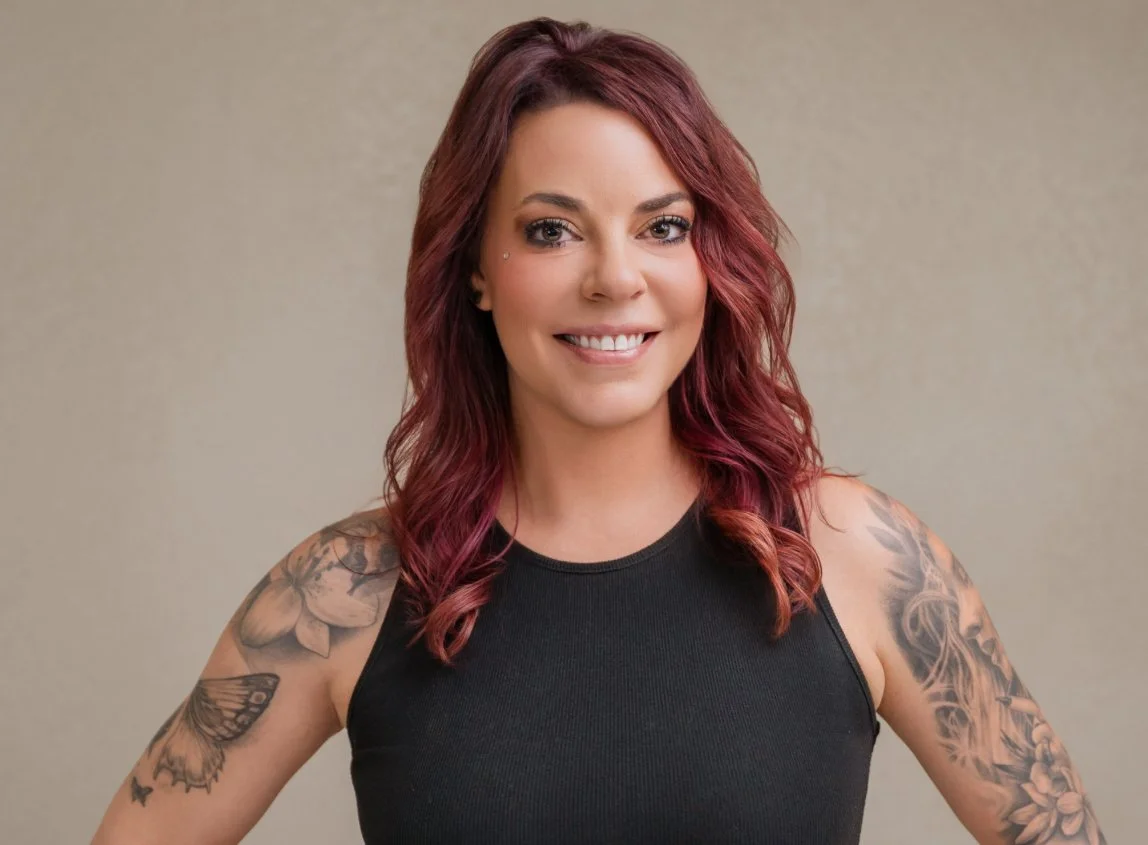 A woman with red hair, tattoos on her arms, and a piercing on her face, wearing a black sleeveless top, smiling at the camera.