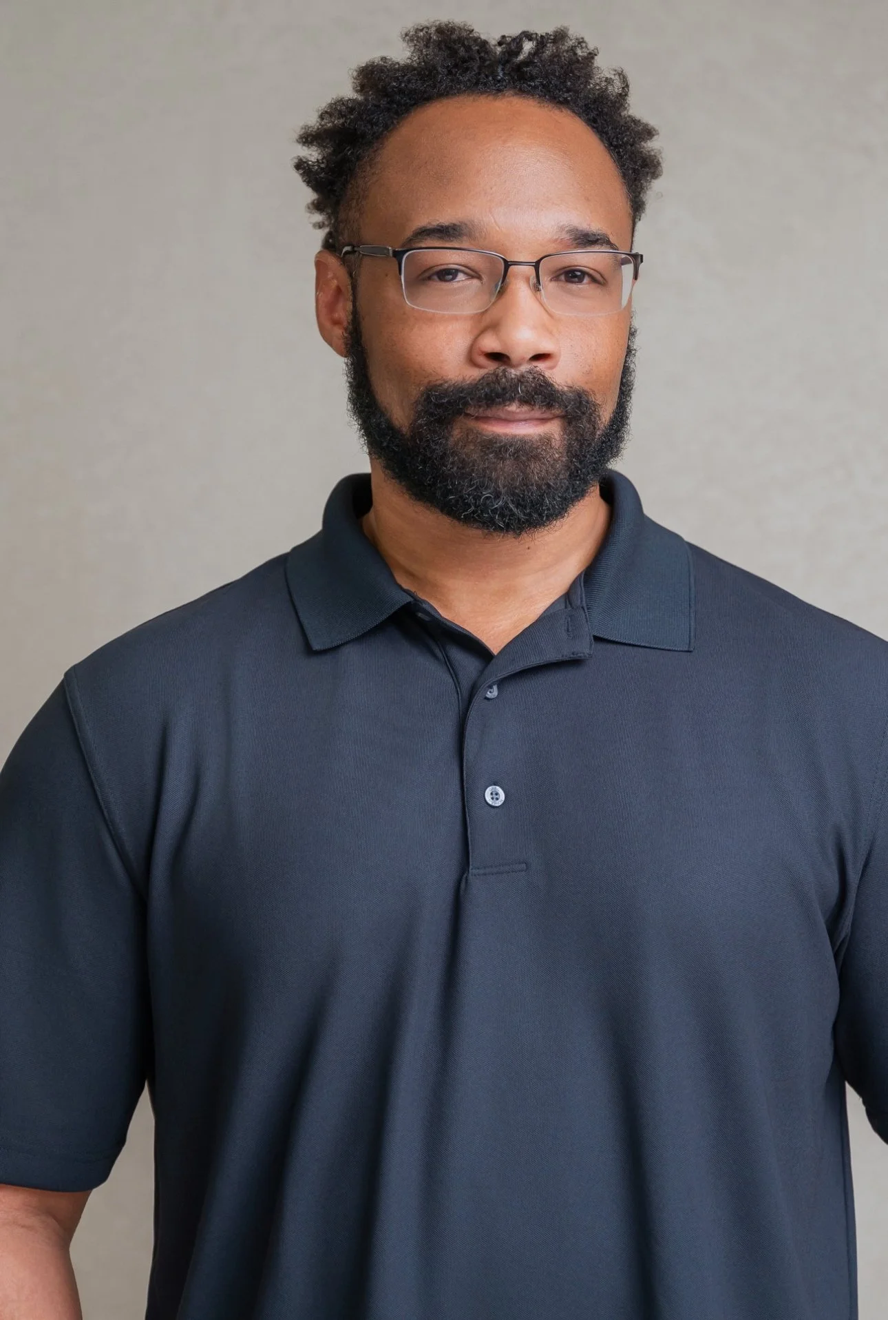A portrait of a man with glasses, a black beard, and mustache, wearing a black polo shirt, standing against a plain light-colored background.