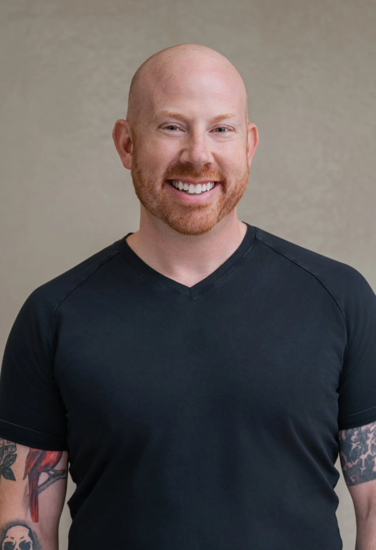 Portrait of a smiling man with a bald head and a red beard, wearing a black V-neck T-shirt, standing against a neutral background.