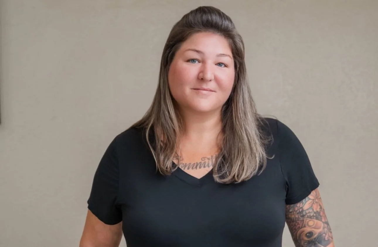 A woman with shoulder-length brown hair wearing a black t-shirt, standing against a plain beige wall.