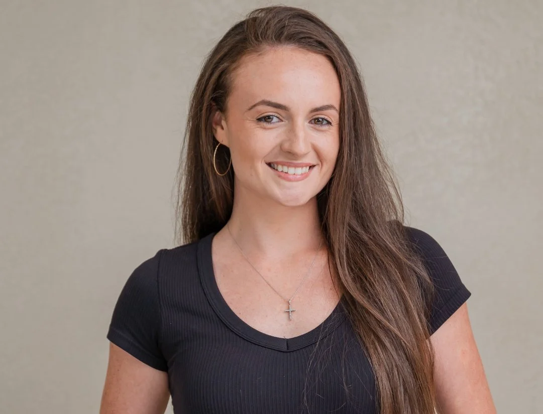 A young woman with long brown hair, wearing a black top, gold hoop earrings, a cross necklace, and smiling.