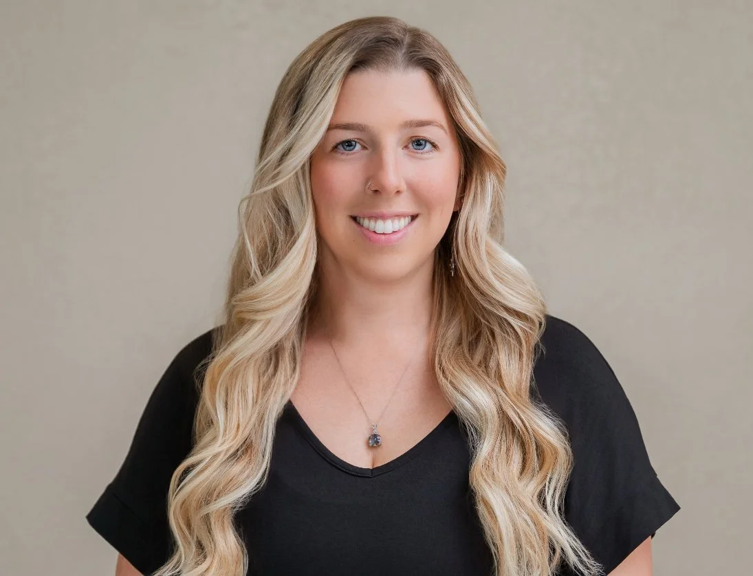 A young woman with long blonde wavy hair, wearing a black top and a necklace, smiling at the camera.
